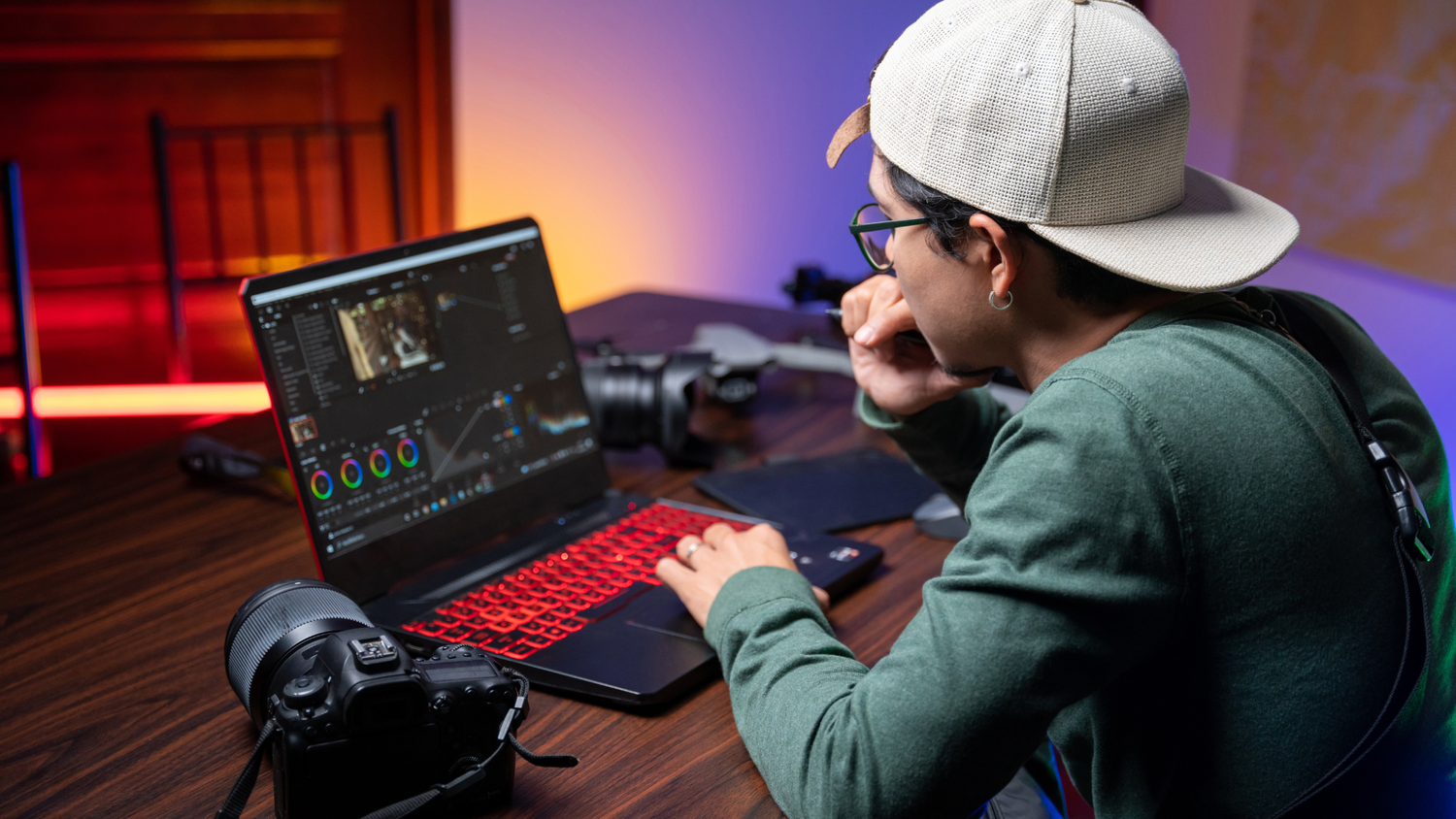 A video editor in a cap reviewing footage on a laptop with color-grading tools open, a camera on the desk beside them and colorful ambient lighting