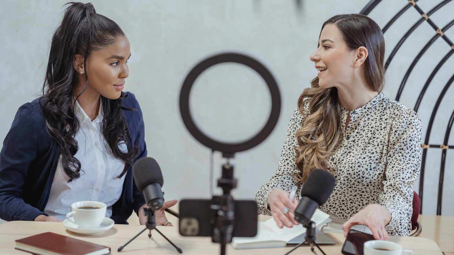 Two women recording a podcast interview at a table with two microphones, a ring light, and a phone mounted on a tripod between them