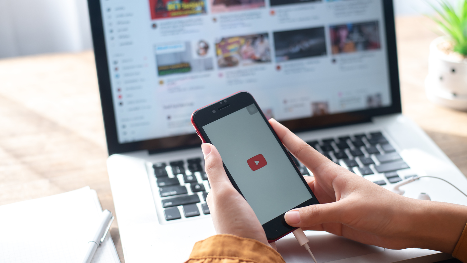 A hand holding a smartphone showing the YouTube play icon in front of a laptop displaying a YouTube feed