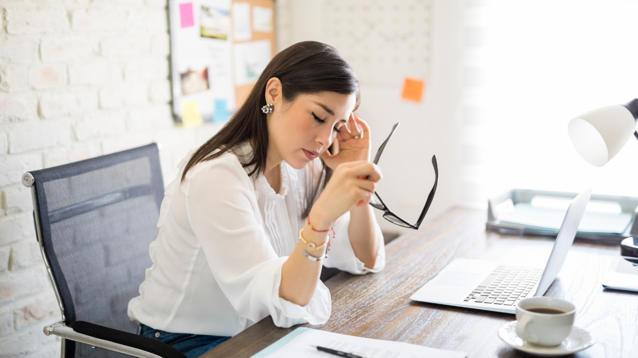 A woman at her desk looking exhausted and stressed, holding her glasses and resting her head against her hand beside a laptop